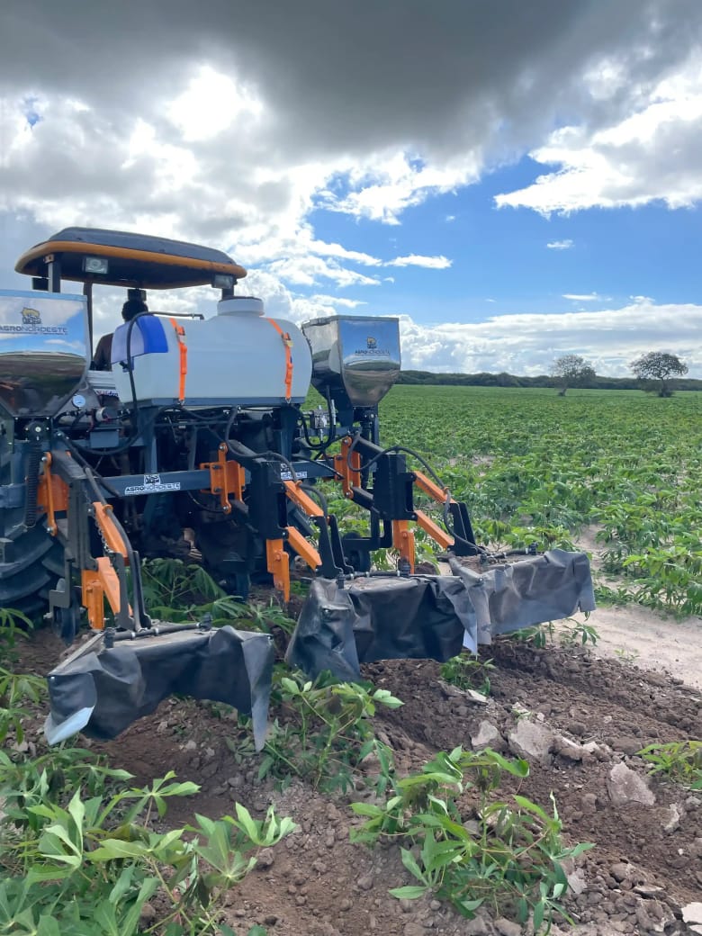 Cultivador Agronordeste acoplado a um trator, com tanque e tremonhas, trabalhando entre linhas de mandioca