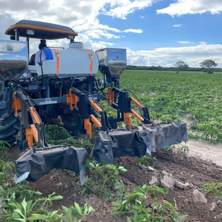 Cultivador Agronordeste acoplado a um trator, com tanque e tremonhas, trabalhando entre linhas de mandioca